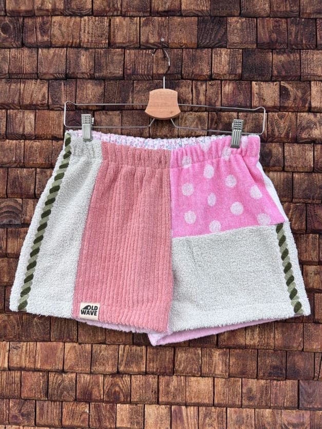 Vorderansicht der Upcycling-Frotteeshorts „Dusty Resort ‘87“ von Old Wave auf einem Kleiderbügel vor einer rustikalen Holzwand. Das Design kombiniert ein linkes Hosenbein in verwaschenem Lachsrosa, das am Saum das Old Wave-Logo trägt, mit einem rechten Hosenbein aus hellgrauem Frottee. Die beiden Hälften werden in der Mitte durch eine vertikale, reich verzierte Orient-Borte mit Musterelementen getrennt.