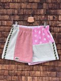 Vorderansicht der Upcycling-Frotteeshorts „Dusty Resort ‘87“ von Old Wave auf einem Kleiderbügel vor einer rustikalen Holzwand. Das Design kombiniert ein linkes Hosenbein in verwaschenem Lachsrosa, das am Saum das Old Wave-Logo trägt, mit einem rechten Hosenbein aus hellgrauem Frottee. Die beiden Hälften werden in der Mitte durch eine vertikale, reich verzierte Orient-Borte mit Musterelementen getrennt.