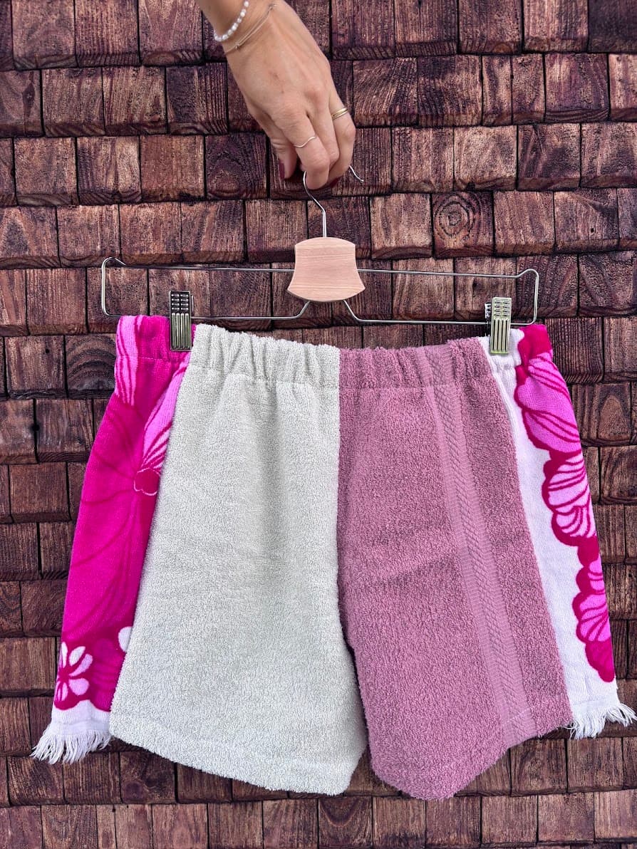Rückansicht der Frottee-Patchwork-Shorts „Flamingo '74“ von Old Wave an einem Kleiderbügel. Diese Perspektive zeigt die asymmetrisch gestaltete Rückseite: Das Gesäß besteht größtenteils aus einem weichen, hellgrau melierten Frotteestoff. Ein großer, quadratischer Patch des lebhaften, blau-weißen Vintage-Fliesenmusters ist unten auf dem rechten Hosenbein platziert, während die linke Außenkante durch einen schmalen Streifen desselben blauen Stoffes akzentuiert wird.