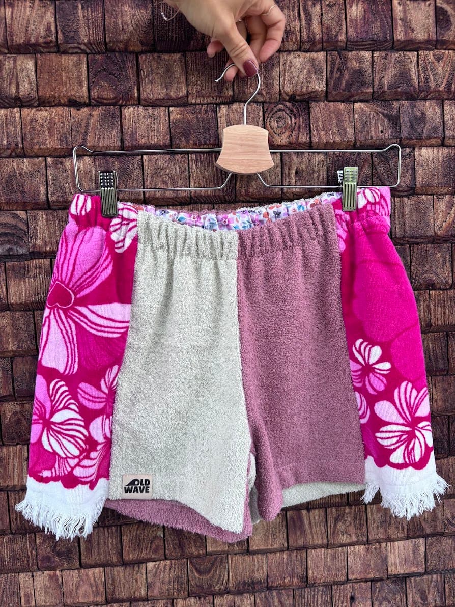 Upcycling-Shorts „Flamingo '74“ von Old Wave auf einem Kleiderbügel vor einer hellen, weißen Zimmertür. Das asymmetrische Patchwork-Design besteht aus upgecycelten Frottee-Handtüchern. Die linke Vorderseite zeigt im oberen Bereich ein markantes, leuchtend blaues Retro-Fliesenmuster mit weißen Ornamenten, das an eine hellgrau melierte Grundfläche grenzt. Das rechte Hosenbein ist vorwiegend in Hellgrau gehalten und schließt an der Außenkante mit einem vertikalen Streifen des blauen Mustermaterials ab.