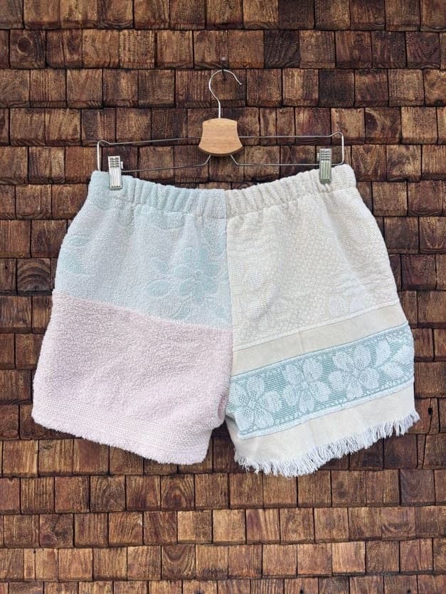 Rückansicht der Upcycling-Frotteeshorts „Fringe Blossom ‘108“ von Old Wave auf einem Kleiderbügel vor einer Holzwand. Das harmonische Patchwork-Muster setzt sich auf der Rückseite fort: Die linke Seite präsentiert große Farbblöcke in Hellblau und softem Altrosa mit gefranstem Saumabschluss, während die rechte Gesäßhälfte komplett in cleanem Weiß gehalten ist.