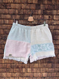 Vorderansicht der Upcycling-Frotteeshorts „Fringe Blossom ‘108“ von Old Wave auf einem Kleiderbügel vor einer rustikalen Holzwand. Das sanfte Pastell-Design besticht durch Fransen am Saum und zeigt ein linkes Hosenbein im dreifarbigen Color-Blocking-Stil aus Hellblau, Altrosa und Weiß samt dem Old Wave-Logo. Das rechte Hosenbein ist in schlichtem Weiß gehalten und mit einem hellblauen Patchwork-Einsatz mit floralem Ornamentmuster verziert.