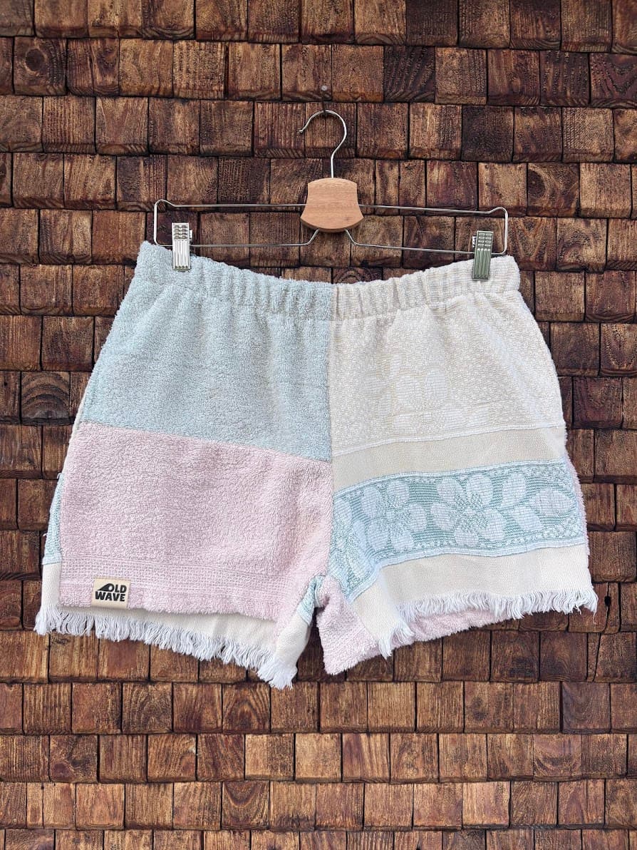 Vorderansicht der Upcycling-Frotteeshorts „Fringe Blossom ‘108“ von Old Wave auf einem Kleiderbügel vor einer rustikalen Holzwand. Das sanfte Pastell-Design besticht durch Fransen am Saum und zeigt ein linkes Hosenbein im dreifarbigen Color-Blocking-Stil aus Hellblau, Altrosa und Weiß samt dem Old Wave-Logo. Das rechte Hosenbein ist in schlichtem Weiß gehalten und mit einem hellblauen Patchwork-Einsatz mit floralem Ornamentmuster verziert.