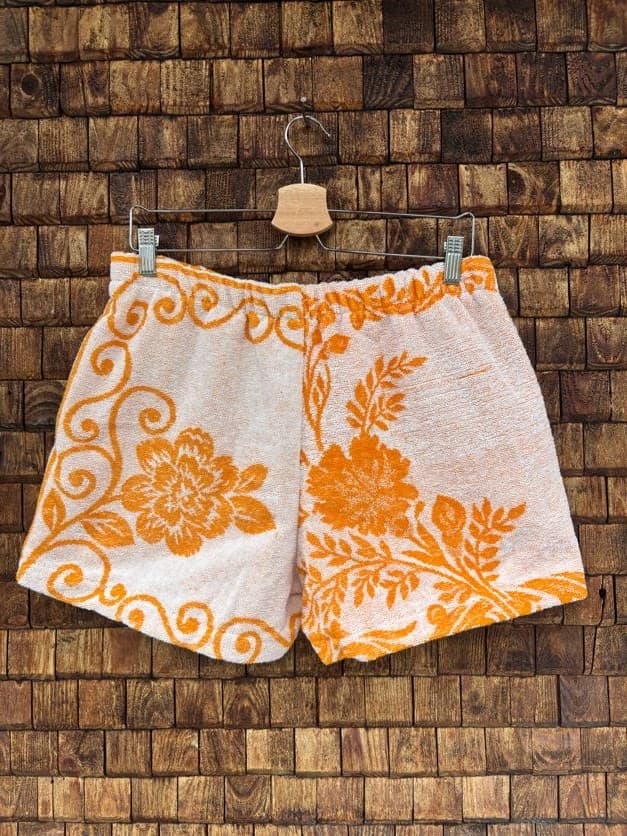 Rückansicht der Upcycling-Frotteeshorts „Giant Bloom ‘70“ von Old Wave auf einem Kleiderbügel vor einer Holzwand. Die Rückseite zeigt ein zweigeteiltes, asymmetrisches Muster-Layout: Die linke Gesäßseite wird von einer großen, kreisrund umrahmten Retro-Blüte geprägt, während die rechte Seite ein filigraneres, dynamisch nach oben verlaufendes florales Blättermotiv in Orange auf Weiß präsentiert.