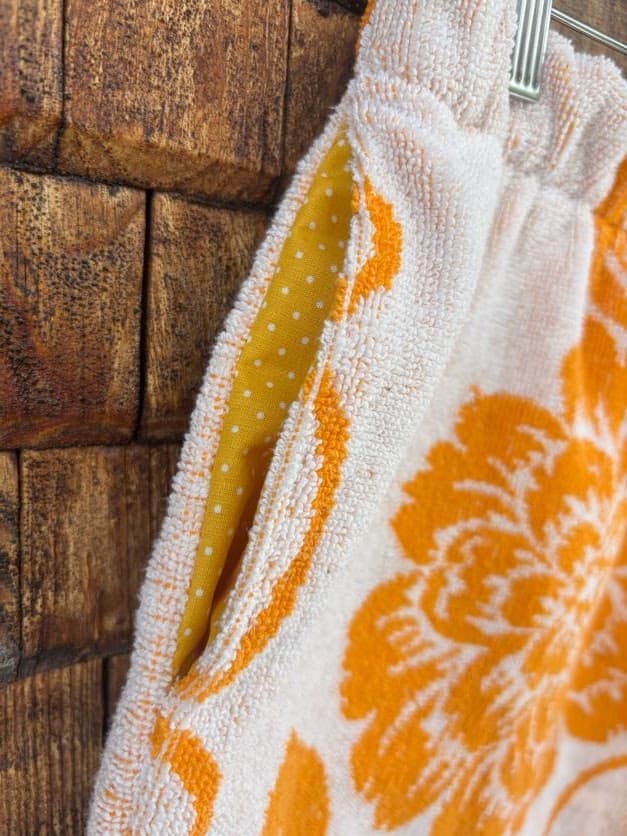 Nahaufnahme der seitlichen Eingrifftasche der „Giant Bloom ‘70“ Frotteeshorts von Old Wave. Das Detail zeigt das farbenfrohe, gelb-weiß gepunktete Innenfutter der Tasche, das einen fröhlichen Kontrast zum weißen Frotteestoff und den großen orangenen Vintage-Blumenmustern bildet.