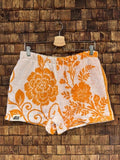 Vorderansicht der Upcycling-Frotteeshorts „Giant Bloom ‘70“ von Old Wave auf einem Kleiderbügel vor einer rustikalen Holzwand. Das sommerliche Retro-Design besticht durch leuchtend orangefarbene, großflächige Blumenmotive und florale Ornamente auf weißem Frotteestoff, abgerundet durch das Old Wave-Logo am linken Saum.