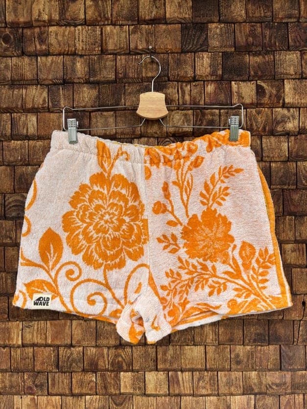 Vorderansicht der Upcycling-Frotteeshorts „Giant Bloom ‘70“ von Old Wave auf einem Kleiderbügel vor einer rustikalen Holzwand. Das sommerliche Retro-Design besticht durch leuchtend orangefarbene, großflächige Blumenmotive und florale Ornamente auf weißem Frotteestoff, abgerundet durch das Old Wave-Logo am linken Saum.