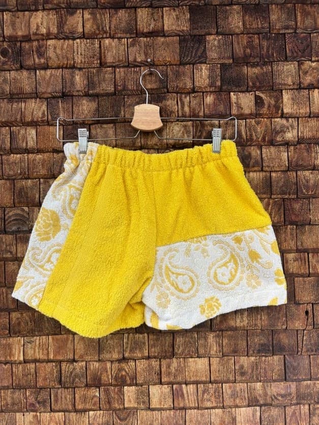 Rückansicht der Upcycling-Frotteeshorts „Golden Paisley ‘71“ von Old Wave auf einem Kleiderbügel vor einer Holzwand. Die Rückseite besticht durch ein asymmetrisches Design: Die linke Gesäßseite besteht überwiegend aus einem unifarbenen, leuchtend gelben Frotteestoff, der an der Außenkante von einem Paisley-Muster flankiert wird. Die rechte Gesäßhälfte ist im oberen Bereich unigelb gehalten und setzt sich im unteren Bereich mit einem großen, kontrastierenden Paisley-Muster-Patch ab.