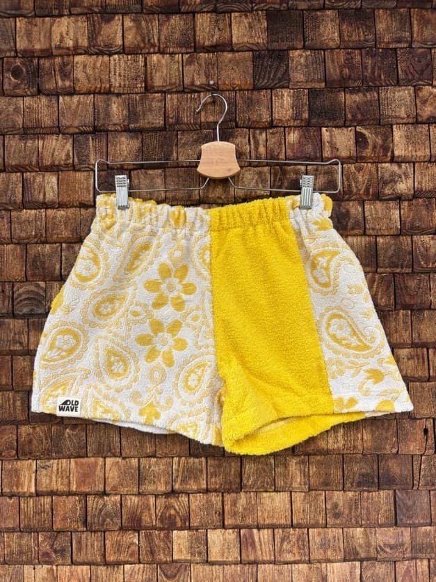 Vorderansicht der Upcycling-Frotteeshorts „Golden Paisley ‘71“ von Old Wave auf einem Kleiderbügel vor einer rustikalen Holzwand. Das sonnengelbe Patchwork-Design zeigt ein linkes Hosenbein mit einem weißen Paisley- und Blumenmuster auf gelbem Grund sowie das Old Wave-Logo am Saum. Das rechte Hosenbein kombiniert ein unifarbenes, strukturiertes gelbes Frottee-Zentrum mit einem seitlich angesetzten, weißen Paisley-Musterstreifen.