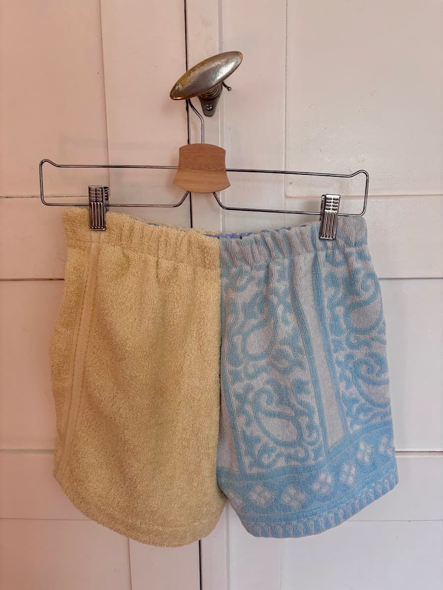 Rückansicht der Frottee-Patchwork-Shorts „Lemon Riviera 61“ von Old Wave. Die Ansicht zeigt die umgekehrte Stoffkombination: Eine Seite besteht aus dem hellblauen Vintage-Handtuchstoff mit geometrischen Mustern und Bordüren, während die andere Seite komplett in schlichtem Zitronengelb gehalten ist.