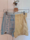 Upcycling-Shorts „Lemon Riviera 61“ von Old Wave an einem Kleiderbügel vor einer weißen Holzwand. Das zweigeteilte Design aus Frottee-Handtüchern kombiniert ein zitronengelbes Hosenbein auf der einen Seite mit einem hellblauen Hosenbein auf der anderen Seite, das mit filigranen, weißen Ornament- und Barockmustern verziert ist.