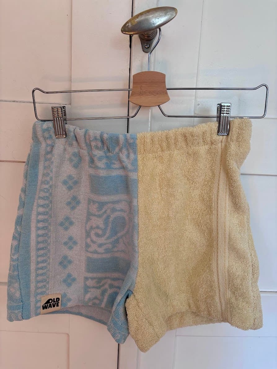 Upcycling-Shorts „Lemon Riviera 61“ von Old Wave an einem Kleiderbügel vor einer weißen Holzwand. Das zweigeteilte Design aus Frottee-Handtüchern kombiniert ein zitronengelbes Hosenbein auf der einen Seite mit einem hellblauen Hosenbein auf der anderen Seite, das mit filigranen, weißen Ornament- und Barockmustern verziert ist.