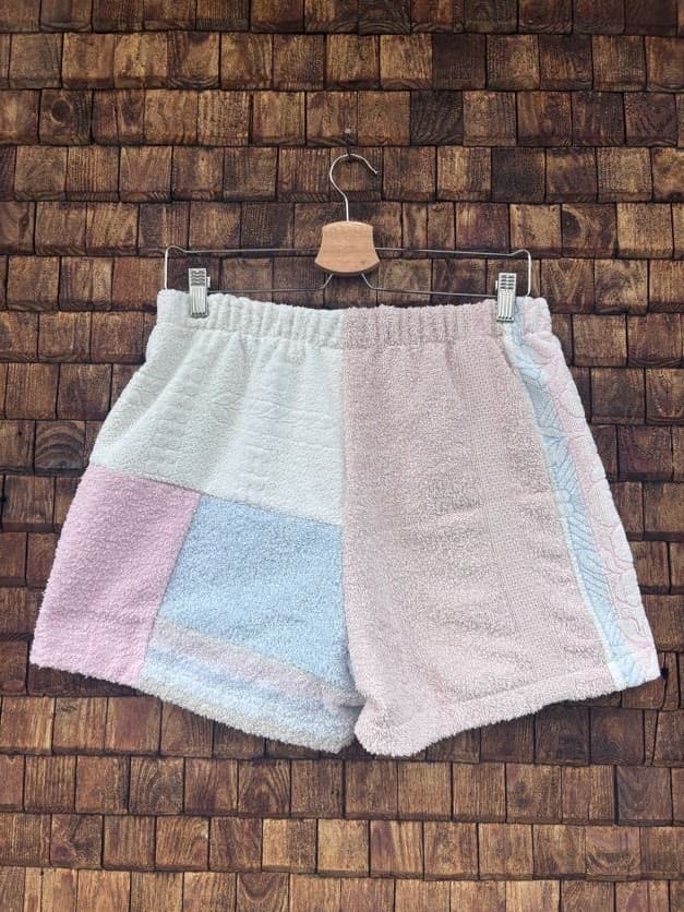 Rückansicht der Upcycling-Frotteeshorts „Lido Ornament ‘99“ von Old Wave auf einem Kleiderbügel vor einer Holzwand. Die Rückseite zeigt ein harmonisches Patchwork-Muster: Ein rechtes Hosenbein in sanftem, meliertem Beige-Rosa trifft auf eine linke Gesäßhälfte, die aus weißen, hellrosa und hellblauen Frottee-Einsätzen asymmetrisch zusammengesetzt ist.