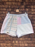 Vorderansicht der Upcycling-Frotteeshorts „Lido Ornament ‘99“ von Old Wave auf einem Kleiderbügel vor einer rustikalen Holzwand. Das pastellfarbene Design präsentiert ein linkes Hosenbein aus weichem Vintage-Frottee mit dezenten, horizontalen Streifenstrukturen und dem Old Wave-Logo am Saum, kombiniert mit einem rechten Hosenbein aus reinem Weiß und farblich abgesetzten Patchwork-Quadraten in Hellrosa und Lichtblau am Bund.