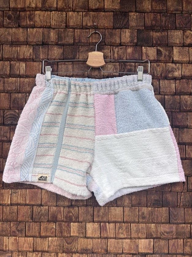 Vorderansicht der Upcycling-Frotteeshorts „Lido Ornament ‘99“ von Old Wave auf einem Kleiderbügel vor einer rustikalen Holzwand. Das pastellfarbene Design präsentiert ein linkes Hosenbein aus weichem Vintage-Frottee mit dezenten, horizontalen Streifenstrukturen und dem Old Wave-Logo am Saum, kombiniert mit einem rechten Hosenbein aus reinem Weiß und farblich abgesetzten Patchwork-Quadraten in Hellrosa und Lichtblau am Bund.