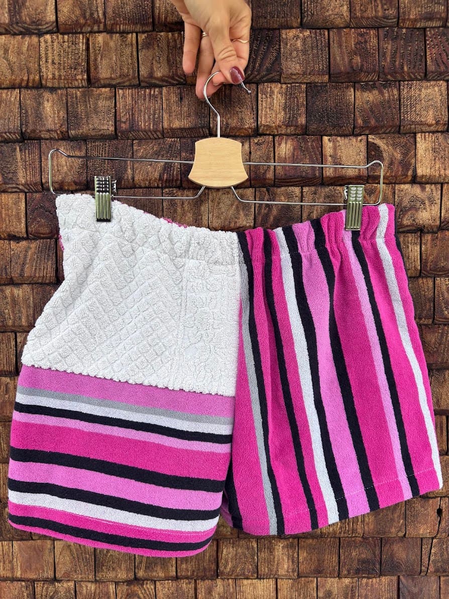 Rückansicht der Frottee-Patchwork-Shorts „Magnolia '81“ von Old Wave an einem Kleiderbügel. Die Rückseite zeigt einen dynamischen Mustermix aus upgecycelten Handtüchern: Das linke Hosenbein wechselt im oberen Bereich zu einer weißen Frotteefläche mit Waffelstruktur über den horizontalen Streifen, während das rechte Hosenbein ein vertikal verlaufendes Streifendesign in den gleichen Pink- und Schwarztönen zeigt.