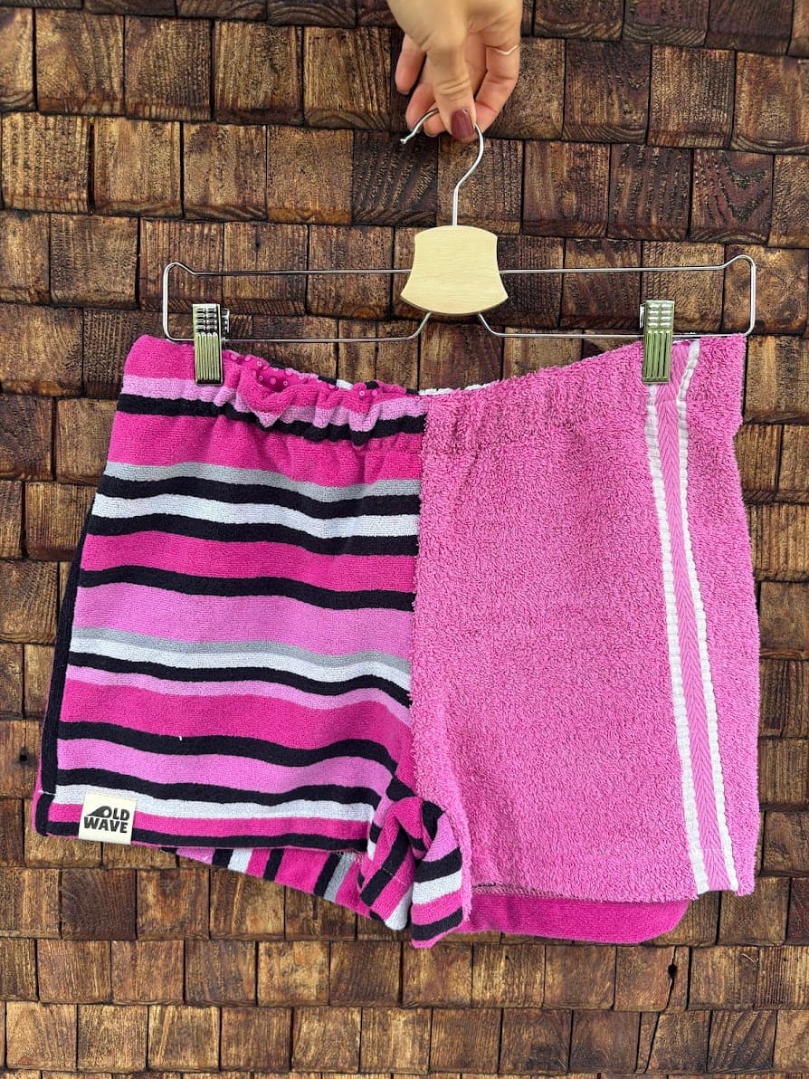 Upcycling-Shorts „Magnolia '81“ von Old Wave auf einem Kleiderbügel vor einer dunklen Holzwand präsentiert. Das asymmetrische Frottee-Design kombiniert ein rechtes Hosenbein aus unifarbenem, kräftigem Rosa mit weißen Sportstreifen am Saum und ein linkes Hosenbein mit einem markanten, horizontalen Retro-Streifenmuster in Pink, Schwarz, Weiß und Grau.