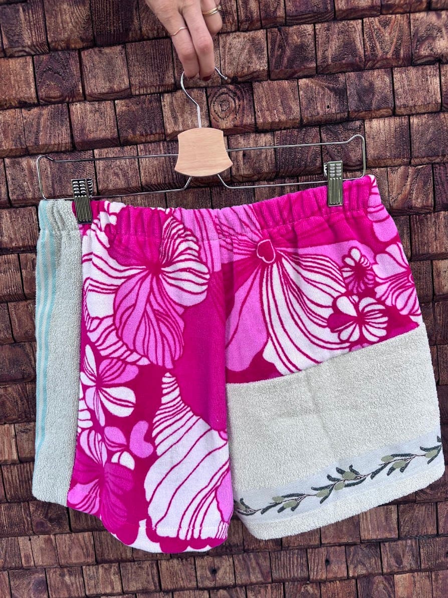 Rückansicht der Frottee-Patchwork-Shorts „Malibu '79“ von Old Wave an einem Kleiderbügel. Diese Perspektive zeigt das asymmetrische Gesäßdesign: Die linke Seite besticht durch ein großflächiges, leuchtend pink-weißes Hibiskus- und Blumenmuster. Die rechte Seite besteht im oberen Teil ebenfalls aus diesem floralen Print, während das untere Drittel des Hosenbeins aus einem mintfarbenen Frotteestoff mit einer feinen, dunklen Ranken-Bordüre am Saum gefertigt ist.