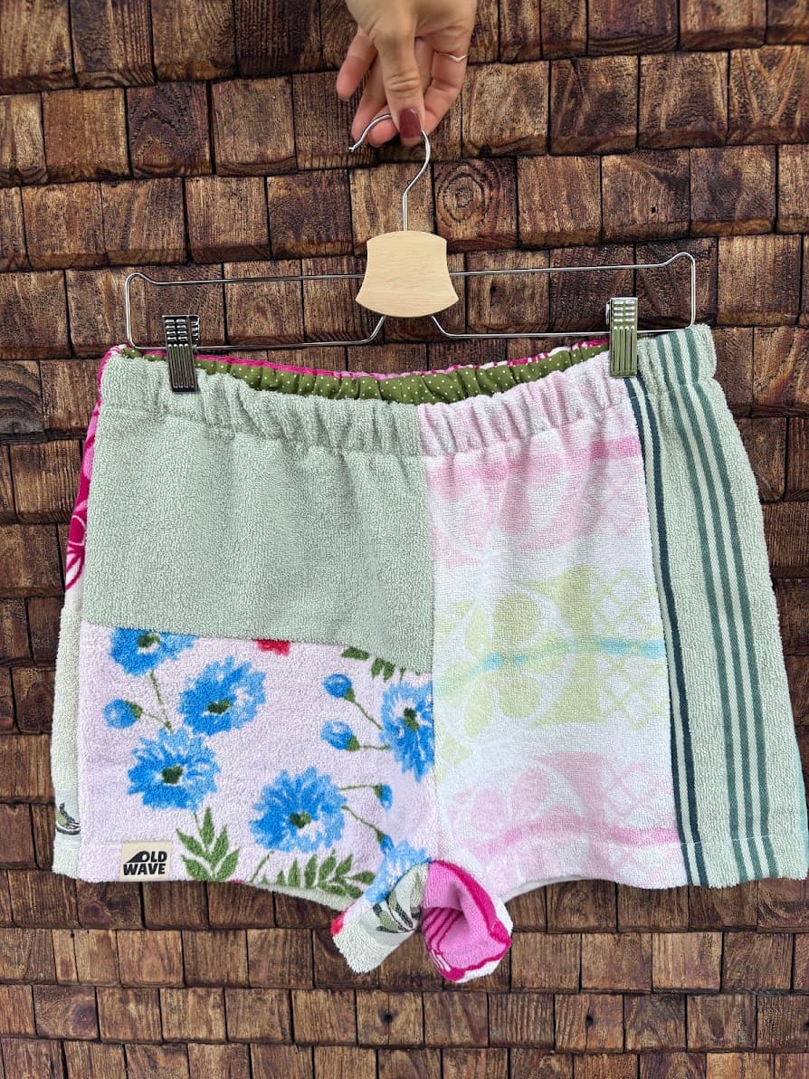 Upcycling-Shorts „Malibu '79“ von Old Wave auf einem Kleiderbügel vor einer dunklen Holzwand. Die Vorderseite zeigt einen kreativen Mustermix aus Frottee-Handtüchern: Das linke Hosenbein kombiniert eine mintgrüne Stoffpartie oben mit einem weiß-blauen Blumenmuster unten, während das rechte Hosenbein aus einem hellrosa-weißen Vintage-Muster besteht, das an der Außenseite von einem olivgrün gestreiften Rand abgeschlossen wird.