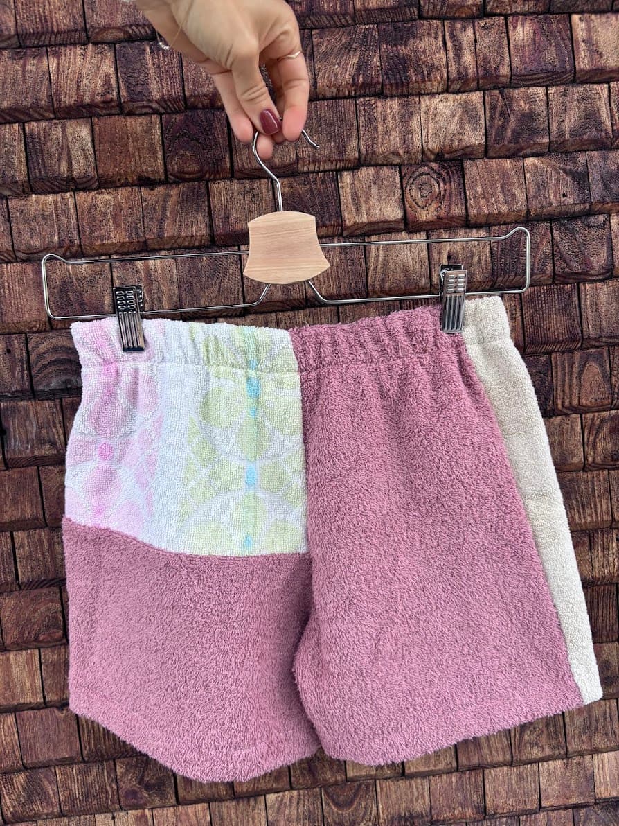 Rückansicht der Frottee-Patchwork-Shorts „Melon '71“ von Old Wave an einem Kleiderbügel. Diese Perspektive zeigt die Rückseite mit einer asymmetrischen Gesäßpartie: Das linke Hosenbein zeigt oben ein dezentes pastellgrün-weißes Kachelmuster und unten matten altrosafarbenen Frotteestoff, während das rechte Hosenbein vorwiegend aus dem altrosafarbenen Stoff besteht und am äußeren Rand von einem beigen Streifen flankiert wird.