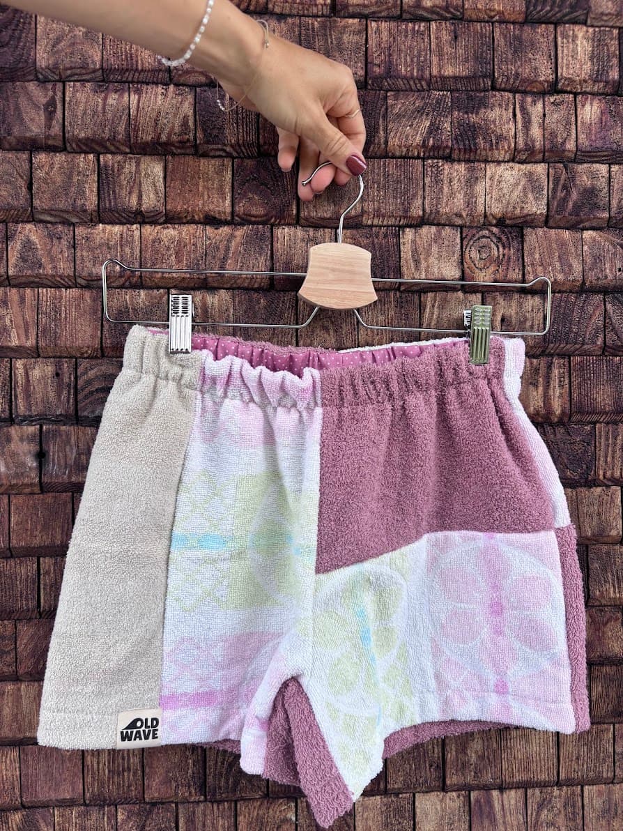 Upcycling-Shorts „Melon '71“ von Old Wave auf einem Kleiderbügel vor einer dunklen Holzwand. Das asymmetrische Patchwork-Design aus Frottee-Handtüchern kombiniert ein linkes Hosenbein aus hellem Beige mit einer gemusterten Mittelfläche in Pastellfarben und ein rechtes Hosenbein in mattem Altrosa, das im unteren Bereich mit einer weißen Patchwork-Fläche samt großen, hellrosa Blütenmotiven abschließt.