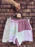 Upcycling-Shorts „Melon '71“ von Old Wave auf einem Kleiderbügel vor einer dunklen Holzwand. Das asymmetrische Patchwork-Design aus Frottee-Handtüchern kombiniert ein linkes Hosenbein aus hellem Beige mit einer gemusterten Mittelfläche in Pastellfarben und ein rechtes Hosenbein in mattem Altrosa, das im unteren Bereich mit einer weißen Patchwork-Fläche samt großen, hellrosa Blütenmotiven abschließt.