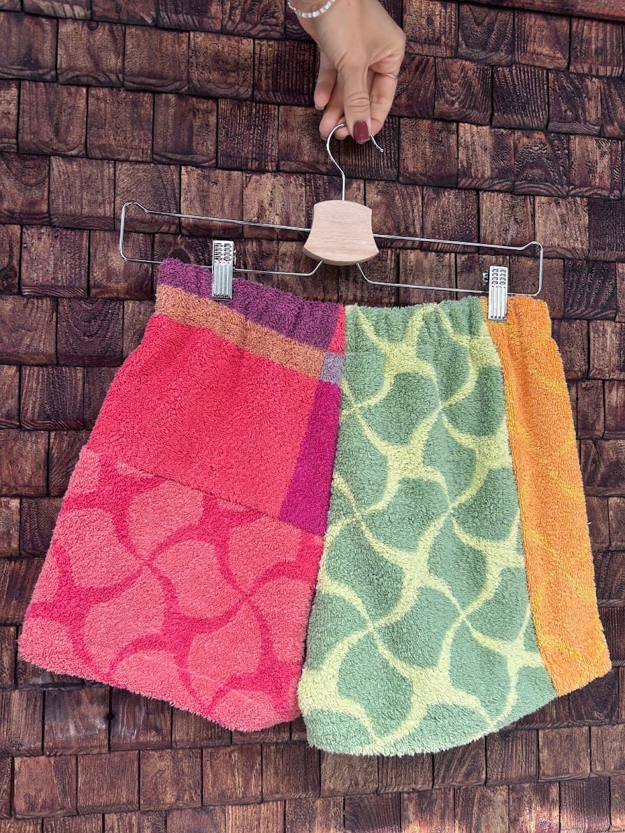 Rückansicht der Upcycling-Frotteeshorts „Mosaic candy stripe '80“ von Old Wave auf einem Kleiderbügel vor einer rustikalen Holzwand. Das farbenfrohe Patchwork-Design kombiniert ein linkes Hosenbein in kräftigem Pink mit einem hellrosa Mosaikmuster und ein rechtes Hosenbein mit mintgrün-gelbem Muster, flankiert von einem orangefarbenen Seitenstreifen.