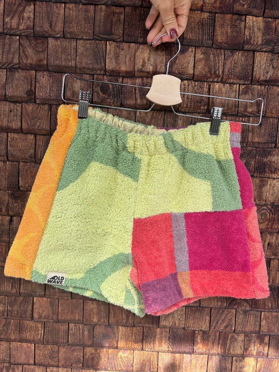 Vorderansicht der Upcycling-Frotteeshorts „Mosaic candy stripe '80“ von Old Wave auf einem Kleiderbügel vor einer Holzwand. Das asymmetrische Design zeigt ein linkes Hosenbein in hellem Gelb-Grün mit einem orangefarbenen Außenstreifen, während das rechte Hosenbein im unteren Bereich durch eine markante, rot-pink karierte Patchwork-Fläche abgeschlossen wird.