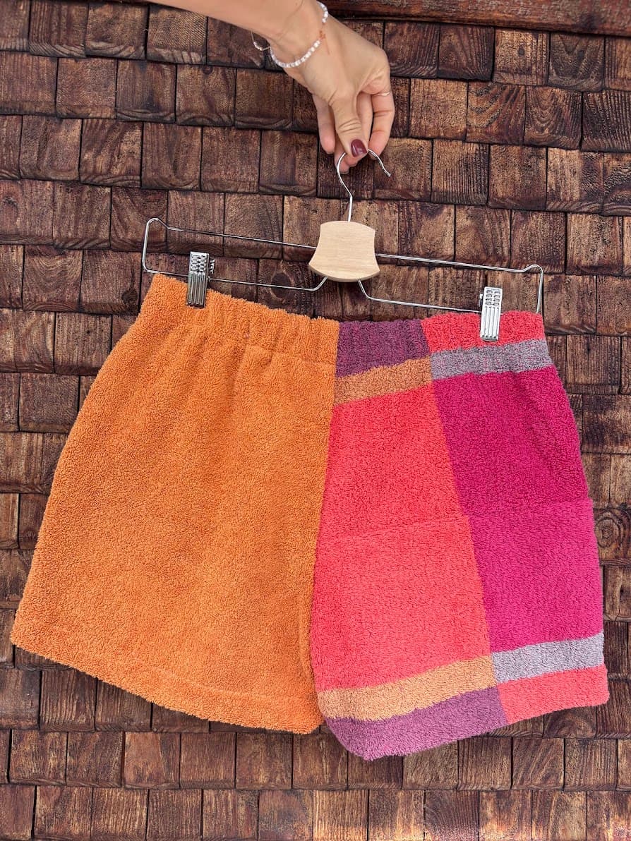 Rückansicht der Upcycling-Frotteeshorts „Mosaic coral bloom '68“ von Old Wave auf einem Kleiderbügel vor einer rustikalen Holzwand. Das asymmetrische Design kombiniert ein linkes Hosenbein aus unifarbenem, kräftig orangefarbenem Vintage-Frotteestoff mit einem rechten Hosenbein im farbenfrohen, großflächigen Karomuster in Rot-, Pink- und Lilatönen.