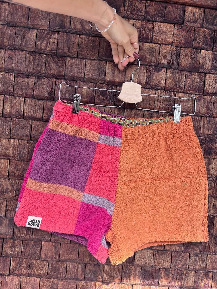 Vorderansicht der Upcycling-Frotteeshorts „Mosaic coral bloom '68“ von Old Wave auf einem Kleiderbügel vor einer Holzwand. Das Design spiegelt die Rückseite: Das linke Hosenbein zeigt das markante Patchwork-Karomuster in Rot, Pink und Lila mit dem eingenähten Old Wave-Logo am Saum, während das rechte Hosenbein schlicht in hellem Orange gehalten ist.