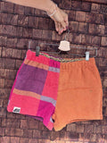 Vorderansicht der Upcycling-Frotteeshorts „Mosaic coral bloom '68“ von Old Wave auf einem Kleiderbügel vor einer Holzwand. Das Design spiegelt die Rückseite: Das linke Hosenbein zeigt das markante Patchwork-Karomuster in Rot, Pink und Lila mit dem eingenähten Old Wave-Logo am Saum, während das rechte Hosenbein schlicht in hellem Orange gehalten ist.