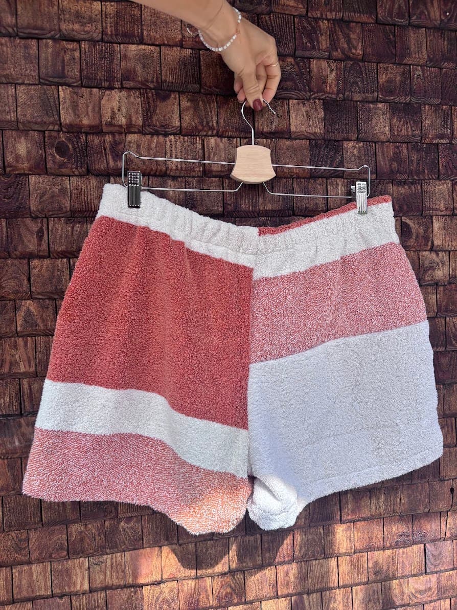Rückansicht der Upcycling-Frotteeshorts „Mosaic coral ripple '70“ von Old Wave auf einem Kleiderbügel vor einer rustikalen Holzwand. Das minimalistische Patchwork-Design zeigt ein linkes Hosenbein in hellem Rotbraun mit einem weißen Querstreifen, kombiniert mit einem rechten Hosenbein in Weiß und einem rotbraun melierten oberen Bereich.