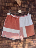 Vorderansicht der Upcycling-Frotteeshorts „Mosaic coral ripple '70“ von Old Wave auf einem Kleiderbügel vor einer Holzwand. Das schlichte Design präsentiert links das rotbraun melierte Hosenbein mit dem weißen Querstreifen und dem eingenähten Old Wave-Logo am Saum, während das rechte Hosenbein vorwiegend in Weiß gehalten ist und rotbraune Patchwork-Akzente setzt.