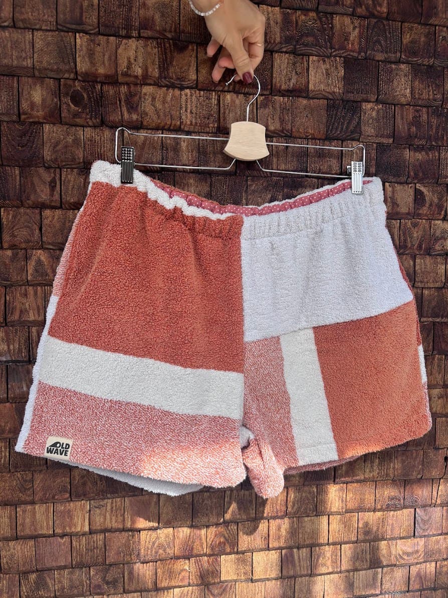 Vorderansicht der Upcycling-Frotteeshorts „Mosaic coral ripple '70“ von Old Wave auf einem Kleiderbügel vor einer Holzwand. Das schlichte Design präsentiert links das rotbraun melierte Hosenbein mit dem weißen Querstreifen und dem eingenähten Old Wave-Logo am Saum, während das rechte Hosenbein vorwiegend in Weiß gehalten ist und rotbraune Patchwork-Akzente setzt.