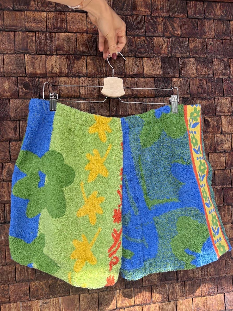 Rückansicht der Upcycling-Frotteeshorts „Mosaic dahlia '72“ von Old Wave auf einem Kleiderbügel vor einer rustikalen Holzwand. Das lebendige Patchwork-Design zeigt ein linkes Hosenbein in Hellgrün mit gelben Retro-Blumenmustern und ein rechtes Hosenbein in kräftigem Blau mit grünen Blüten- und Palmenmotiven, abgerundet durch eine farbige Vintage-Bordüre am äußeren Rand.