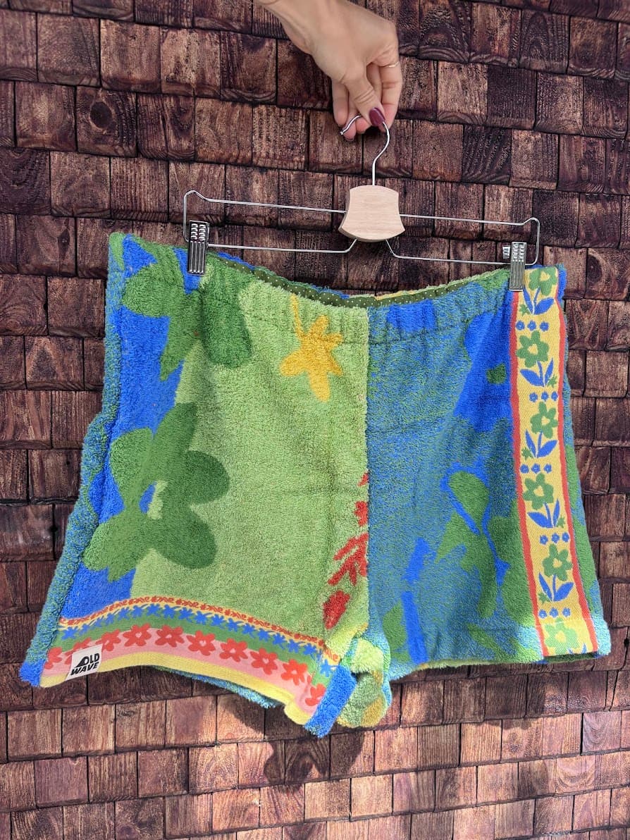 Vorderansicht der Upcycling-Frotteeshorts „Mosaic dahlia '72“ von Old Wave auf einem Kleiderbügel vor einer Holzwand. Das asymmetrische tropische Muster präsentiert links das blaue Frotteematerial mit dem eingenähten Old Wave-Logo am Saum, kombiniert mit der hellgrünen Stoffhälfte auf der rechten Seite und einer auffälligen, bunt gemusterten Bordüre entlang des Hosenbeins.