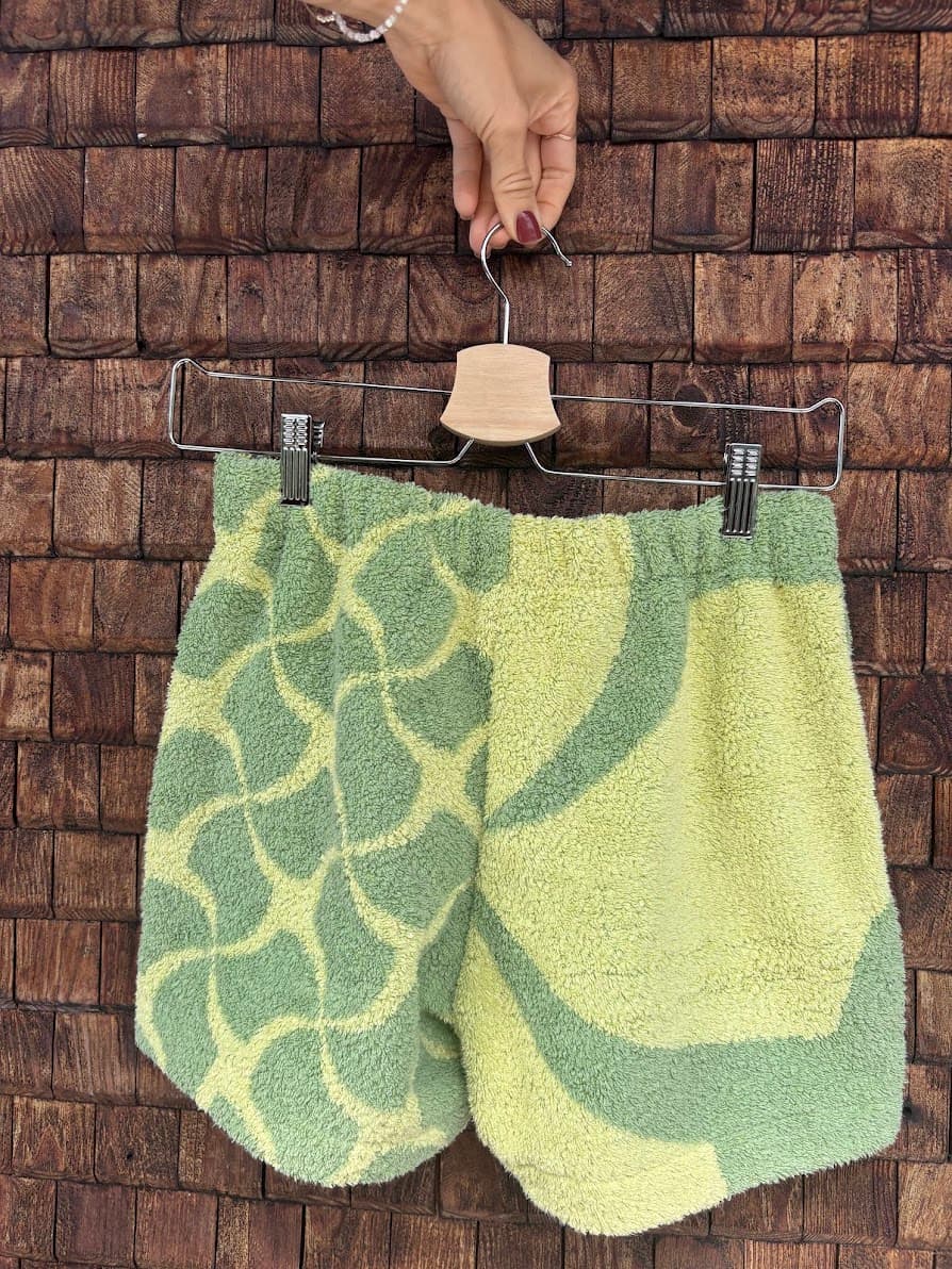 Rückansicht der Upcycling-Frotteeshorts „Mosaic green '74“ von Old Wave auf einem Kleiderbügel vor einer rustikalen Holzwand. Das asymmetrische Patchwork-Design aus Vintage-Handtüchern zeigt auf dem linken Hosenbein ein kleinteiliges Mosaikmuster in Mintgrün und Gelb, während das rechte Hosenbein ein großflächigeres, geschwungenes Muster in denselben Farben aufweist.
