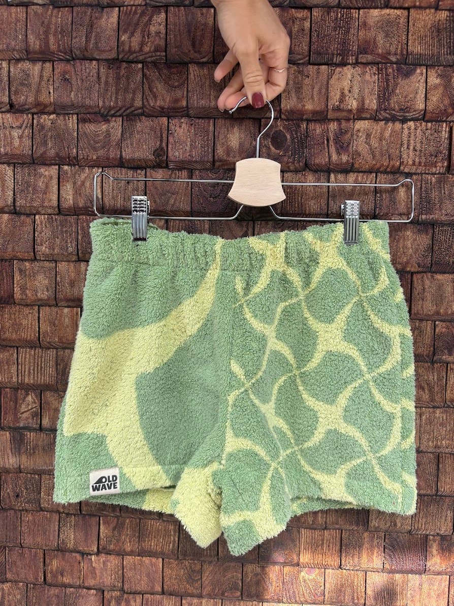 Vorderansicht der Upcycling-Frotteeshorts „Mosaic green '74“ von Old Wave auf einem Kleiderbügel vor einer Holzwand. Das nachhaltige Design vertauscht die Musterung asymmetrisch zur Rückseite: Das linke Bein zeigt das großflächige, geschwungene Wellenmuster in Mintgrün und Gelb mit dem eingenähten Old Wave-Logo am Saum, während das rechte Hosenbein das kleinteiligere Mosaikmuster präsentiert.