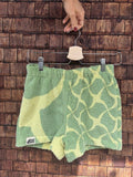 Vorderansicht der Upcycling-Frotteeshorts „Mosaic green '74“ von Old Wave auf einem Kleiderbügel vor einer Holzwand. Das nachhaltige Design vertauscht die Musterung asymmetrisch zur Rückseite: Das linke Bein zeigt das großflächige, geschwungene Wellenmuster in Mintgrün und Gelb mit dem eingenähten Old Wave-Logo am Saum, während das rechte Hosenbein das kleinteiligere Mosaikmuster präsentiert.