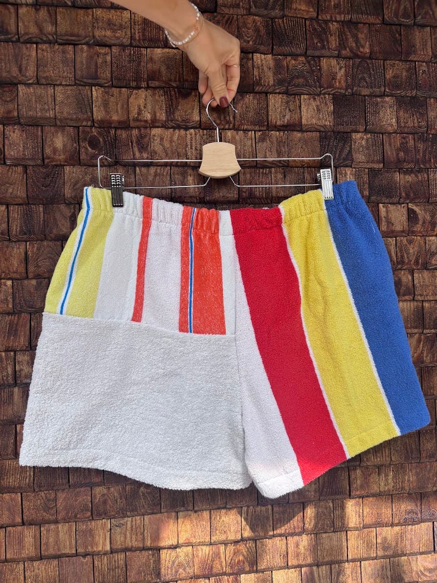 Rückansicht der Upcycling-Frotteeshorts „Mosaic horizon '82“ von Old Wave auf einem Kleiderbügel vor einer rustikalen Holzwand. Das sportliche Patchwork-Design kombiniert ein linkes Hosenbein aus weißem Frottee mit bunten Streifenakzenten im oberen Bereich und ein rechtes Hosenbein mit breiten Längsstreifen in Rot, Gelb und Blau.