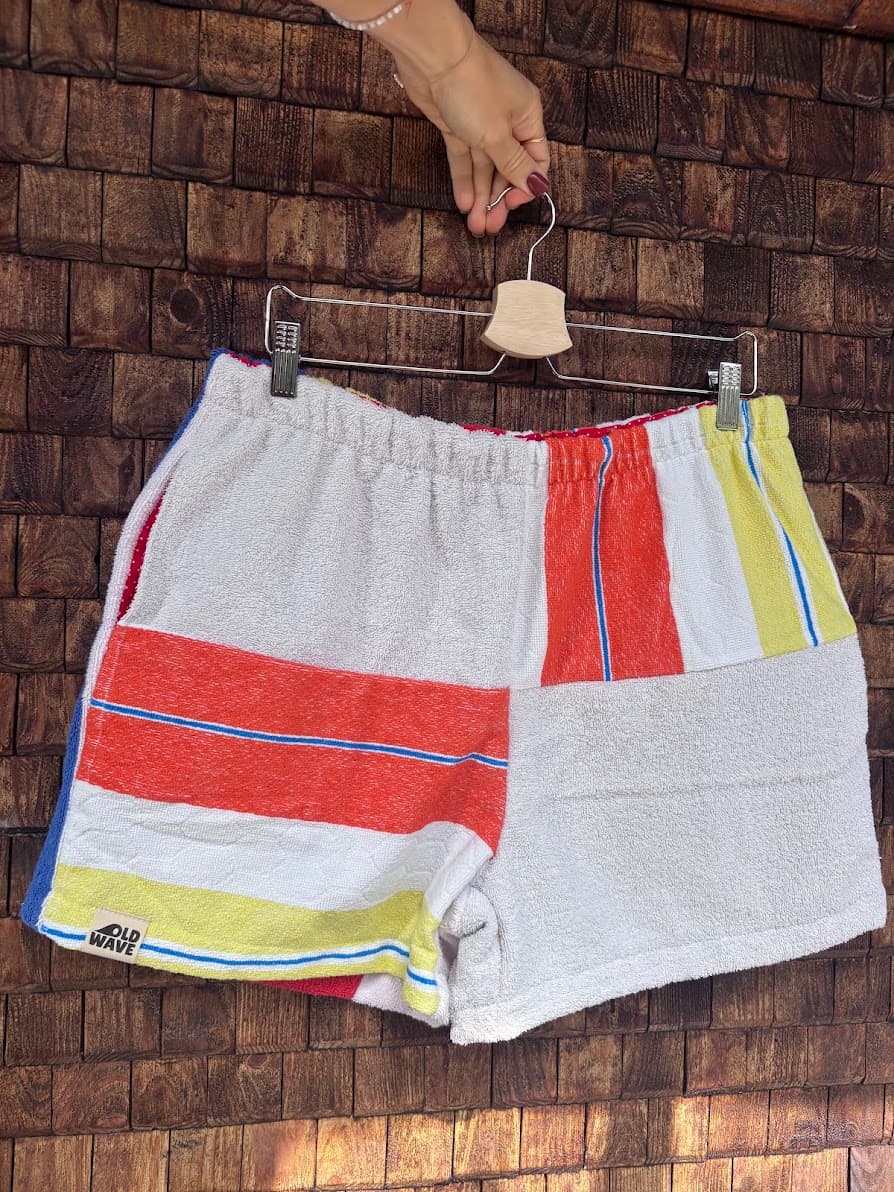 Vorderansicht der Upcycling-Frotteeshorts „Mosaic horizon '82“ von Old Wave auf einem Kleiderbügel vor einer Holzwand. Das asymmetrische Design zeigt links breite Querstreifen in Rot, Gelb und Blau mit dem eingenähten Old Wave-Logo am Saum, während das rechte Hosenbein vorwiegend aus weißem Vintage-Frotteestoff mit farbigen Akzenten am Bund besteht.