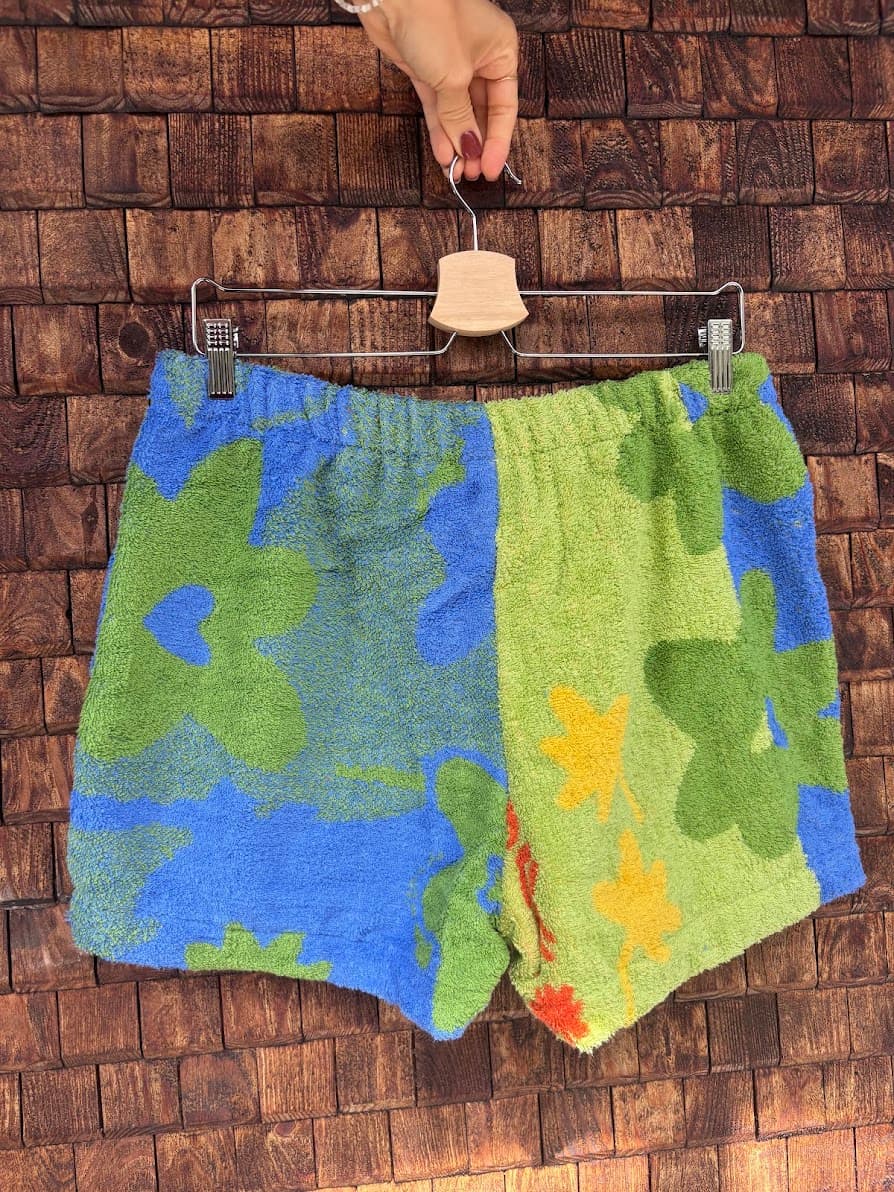 Rückansicht der Upcycling-Frotteeshorts „Mosaic jungle '85“ von Old Wave auf einem Kleiderbügel vor einer rustikalen Holzwand. Das farbenfrohe Patchwork-Design zeigt links ein blaues Muster mit großen grünen Blüten, während das rechte Hosenbein aus hellgrünem Frotteestoff mit gelben und grünen Blattelemeten besteht.