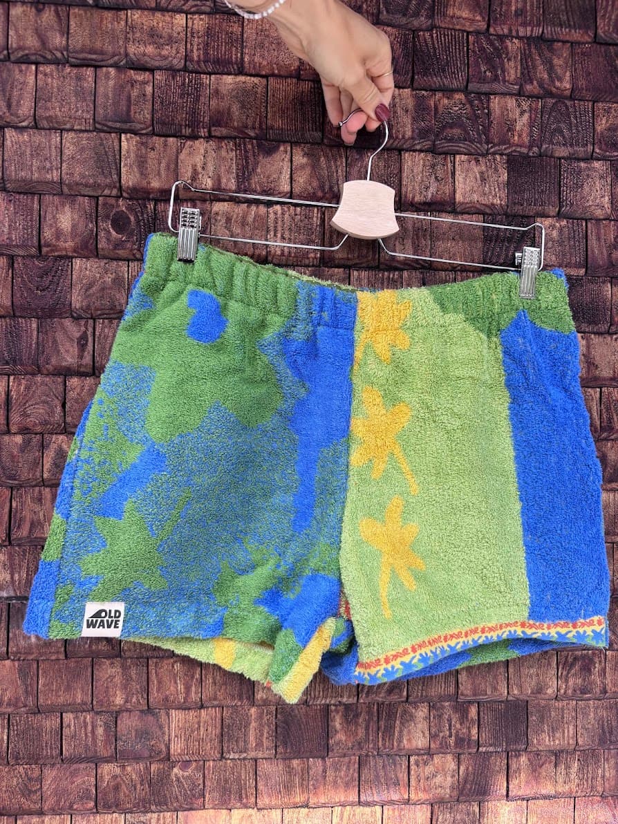 Vorderansicht der Upcycling-Frotteeshorts „Mosaic jungle '85“ von Old Wave auf einem Kleiderbügel vor einer Holzwand. Das asymmetrische tropische Design vertauscht die Musterung: Links ist das blaue Frotteematerial mit grünen Blüten und dem eingenähten Old Wave-Logo am Saum zu sehen, rechts das hellgrüne Muster mit gelben Akzenten und einem blauen Seitenstreifen.