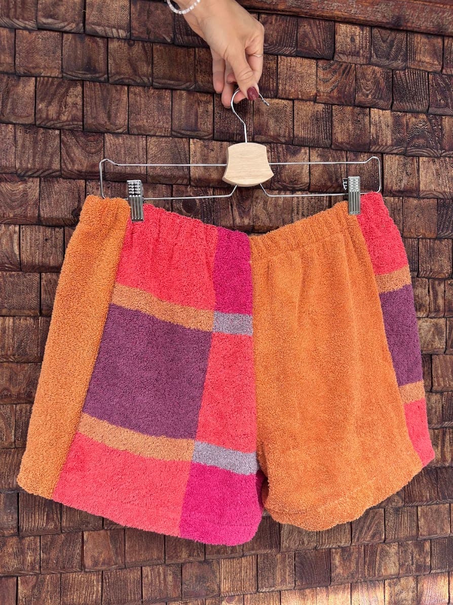 Rückansicht der Upcycling-Frotteeshorts „Mosaic plum wave '77“ von Old Wave auf einem Kleiderbügel vor einer rustikalen Holzwand. Das asymmetrische Patchwork-Design zeigt links ein markantes Karomuster in Orange, Lila und Pink, während das rechte Hosenbein schlicht aus unifarbenem, hellem Orange besteht.