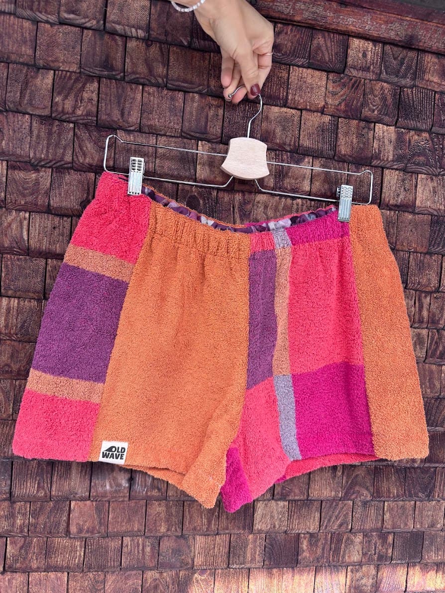Vorderansicht der Upcycling-Frotteeshorts „Mosaic plum wave '77“ von Old Wave auf einem Kleiderbügel vor einer Holzwand. Das gespiegelte Design zeigt hier das linke Hosenbein in unifarbenem Orange mit dem eingenähten Old Wave-Logo am Saum, kombiniert mit einem rechten Hosenbein im farbenfrohen Karomuster aus Orange-, Lila- und Pinktönen.
