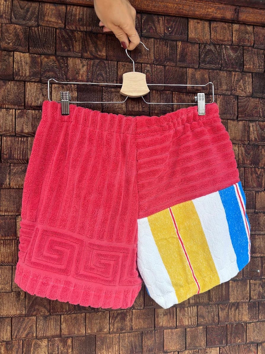 Rückansicht der Upcycling-Frotteeshorts „Mosaic retro floral '76“ von Old Wave auf einem Kleiderbügel vor einer rustikalen Holzwand. Das asymmetrische Design zeigt ein linkes Hosenbein in kräftigem Rot mit einer eingewebten griechischen Mäander-Bordüre am Saum, kombiniert mit einem rechten Hosenbein, das im unteren Bereich Querstreifen in Gelb, Weiß und Blau aufweist.