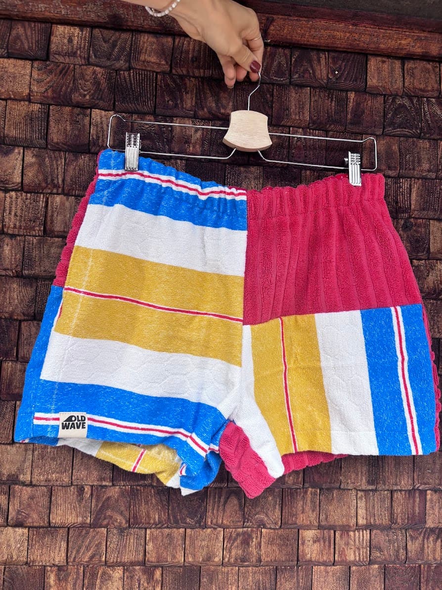 Vorderansicht der Upcycling-Frotteeshorts „Mosaic retro floral '76“ von Old Wave auf einem Kleiderbügel vor einer Holzwand. Das auffällige Patchwork-Muster zeigt auf der linken Seite breite Querstreifen in Gelb, Weiß und Blau mit dem eingenähten Old Wave-Logo am Saum, während das rechte Bein aus rotem Frotteestoff besteht und am äußeren Rand gestreifte Akzente setzt.