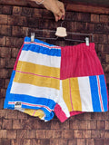 Vorderansicht der Upcycling-Frotteeshorts „Mosaic retro floral '76“ von Old Wave auf einem Kleiderbügel vor einer Holzwand. Das auffällige Patchwork-Muster zeigt auf der linken Seite breite Querstreifen in Gelb, Weiß und Blau mit dem eingenähten Old Wave-Logo am Saum, während das rechte Bein aus rotem Frotteestoff besteht und am äußeren Rand gestreifte Akzente setzt.