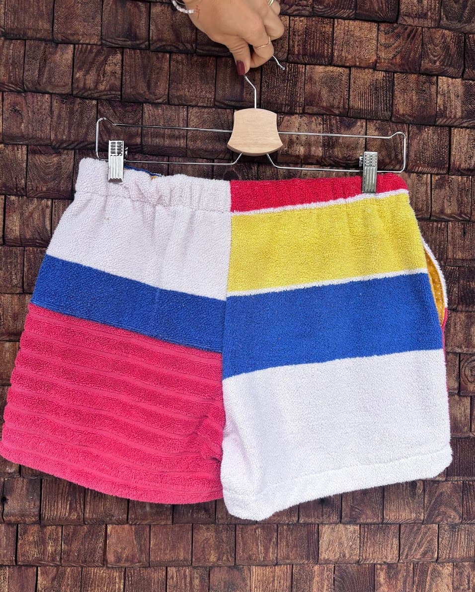 Rückansicht der Upcycling-Frotteeshorts „Mosaic sea breeze '81“ von Old Wave auf einem Kleiderbügel vor einer rustikalen Holzwand. Das maritime Patchwork-Design zeigt links eine Aufteilung in Weiß, Blau und strukturiertes Pink, während das rechte Hosenbein breite Querstreifen in Gelb, Blau und Weiß aufweist.
