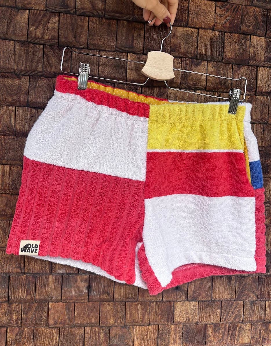 Vorderansicht der Upcycling-Frotteeshorts „Mosaic sea breeze '81“ von Old Wave auf einem Kleiderbügel vor einer Holzwand. Das asymmetrische Design zeigt ein linkes Hosenbein in Weiß und kräftigem Rot mit dem eingenähten Old Wave-Logo am Saum, kombiniert mit einem rechten Hosenbein in Gelb, Rot und Weiß.